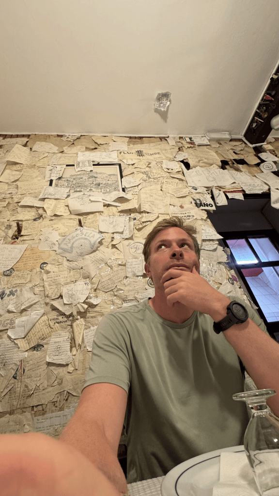 A young man in a green shirt sits at a table, thoughtfully resting his chin on his hand. Behind him is a wall covered with various handwritten notes and papers.