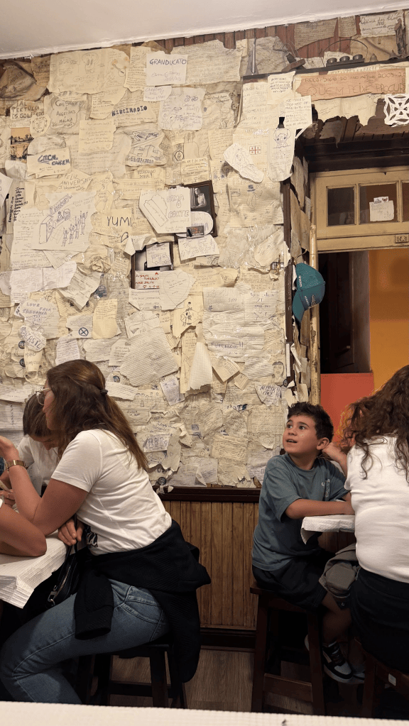 Interior of a restaurant featuring a wall covered with notes and drawings, with people seated at tables, showcasing a cozy and lively atmosphere.
