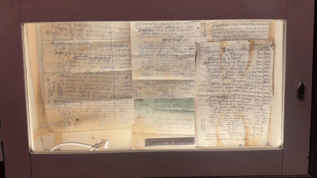 A collection of handwritten menus and notes displayed in a glass case, featuring various food items and prices in Spanish.